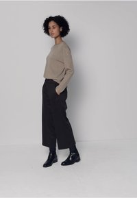 Suéter de punto beige y pantalones negros de pierna ancha, combinados con botines negros. La modelo está de pie frente a un fondo gris claro, con las manos en los bolsillos.