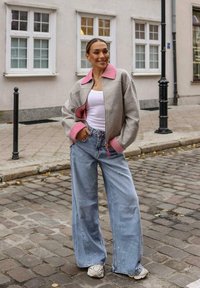 Šedá a růžová bunda na zip s širokými rukávy, nošená přes bílý tílko, v kombinaci se světle modrými volnými džíny a bílými teniskami. Pozadí s dlažebními kameny.