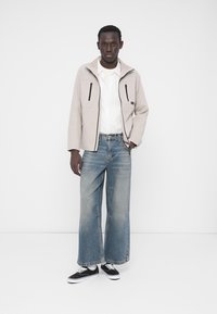 Veste beige clair zippée avec deux poches poitrine portée sur une chemise blanche, associée à un jean bleu délavé à jambes larges et des baskets noires.