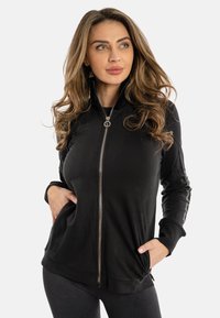Giacca nera con zip realizzata in tessuto morbido, con colletto alto, tasche laterali e dettagli decorativi lungo le maniche.