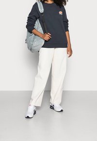 Felpa blu navy scuro con polsini elasticizzati, pantaloni bianchi a gamba larga e zaino in velluto a coste azzurro chiaro; sneakers bianche e blu navy completano il look.