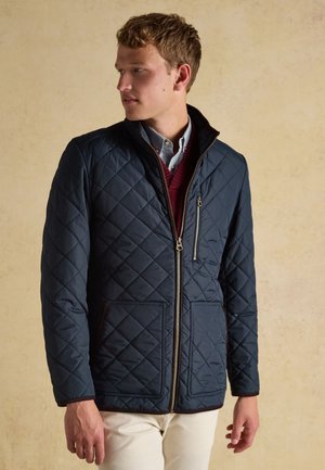 Young man wearing a navy quilted jacket over a striped shirt and burgundy sweater, looking to the left against a plain beige background.