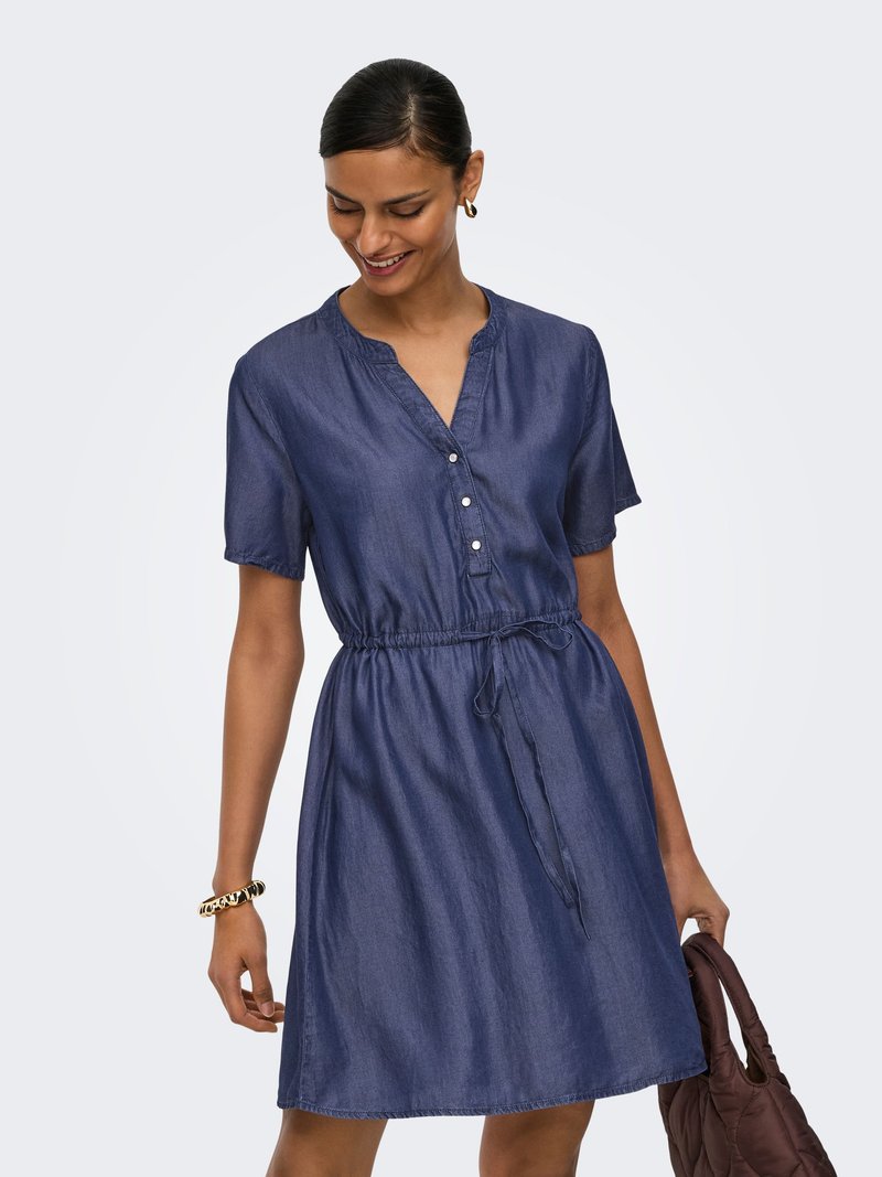 Femme portant une robe bleu marine à manches courtes, boutonnée, avec une taille resserrée par un cordon, tenant un sac à main matelassé marron dans sa main gauche.