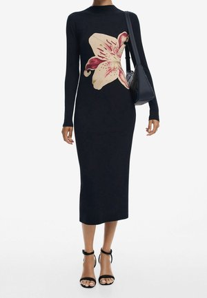 Kobieta w długiej, czarnej dopasowanej sukience z dużym różowo-białym kwiatowym nadrukiem, czarna torebka i czarne szpilki z zapięciem wokół kostki.