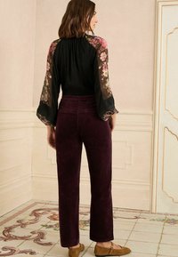 Čierny top s kvetinovými priehľadnými rukávmi, skombinovaný s bordovými kordovým nohavicami. Ploské hnedé topánky dopĺňajú outfit, spolu s dekoratívnym vzorom na podlahe.