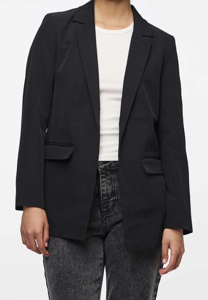 Personne portant un blazer noir sur une chemise blanche côtelée associée à un jean gris foncé taille haute, debout devant un fond uni.