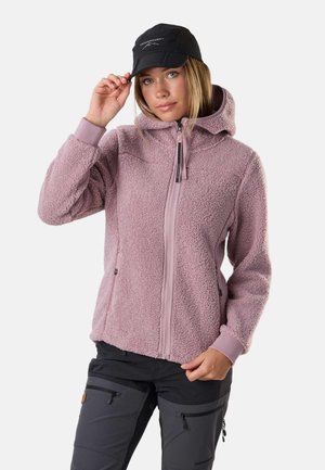 Kvinde iført en lyse lilla fleecejakke med hætte, sort kasket og sorte udendørsbukser, justerer kasketten med den ene hånd.
