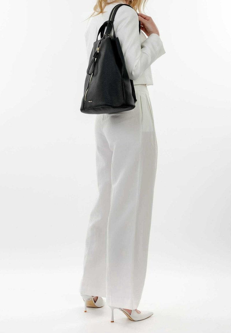 Zwarte leren rugzak met een gestructureerde afwerking, voorzien van een gouden rits en een contrasterende klep, gedragen over een witte outfit.