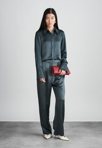 Sötétzöld selyem ing és az ahhoz illő, lazán szabott nadrág; piros clutch a kézben; krémszínű, hegyes orrú magassarkú cipő; sima textúra.