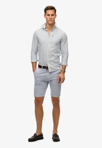 Camicia button-up a righe blu chiaro con maniche arrotolabili, abbinata a pantaloni corti blu chiaro. Una cintura marrone e mocassini marrone scuro completano l'outfit.