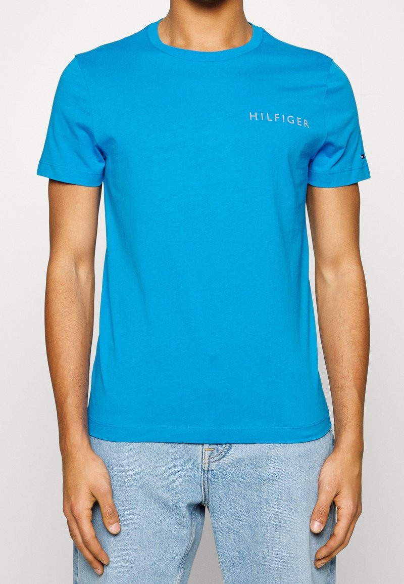 Maglietta in cotone blu con maniche corte, colletto a girocollo e logo "HILFIGER" in bianco sul lato sinistro del petto. Abbinata a jeans azzurri chiari.