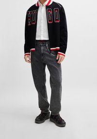 Zwarte bomberjack met rode en witte accenten, voorzien van de tekst "HUGO". Gecombineerd met grijze denim broek en zwarte sneakers.