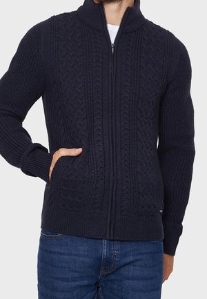 Mann trägt einen marineblauen Strickpullover mit Reißverschluss, hohem Kragen und vorderen Taschen, kombiniert mit blauen Jeans.