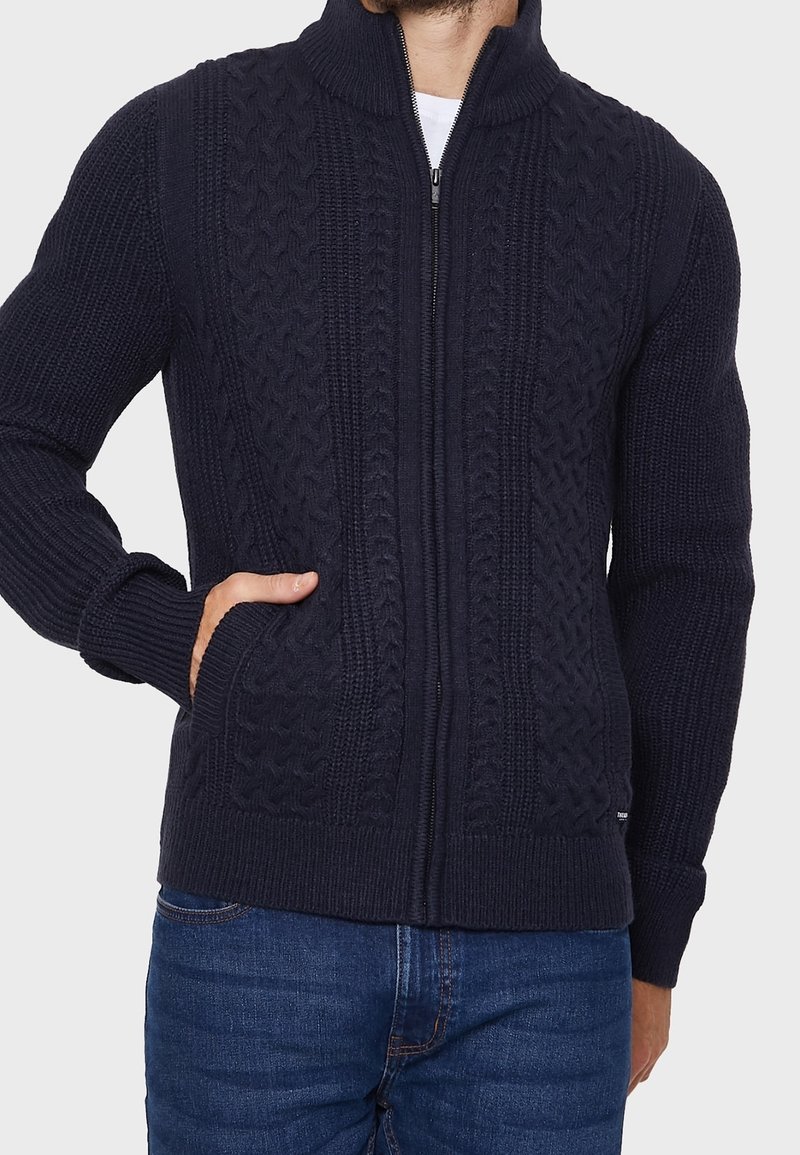 Homme portant un pull en maille torsadée bleu marine zippé avec col montant et poches avant, associé à un jean bleu.
