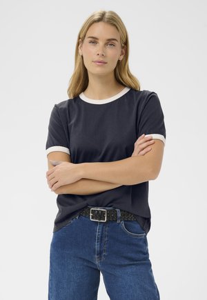 Jonge vrouw met blond haar, gekleed in een zwart T-shirt met witte bies, een blauwe spijkerbroek en een zwarte gevlochten riem, met gekruiste armen.