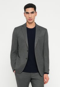 Man in een grijze twee-knops colbert en bijpassende broek boven een zwart T-shirt met ronde hals, staand met één hand in zijn zak.
