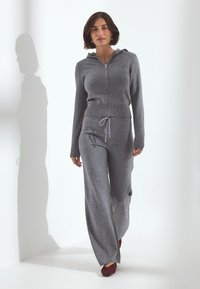 Donna che cammina indossando una felpa grigia con zip e pantaloni a gamba larga abbinati, completati da scarpe rosso scuro, su sfondo bianco uniforme.