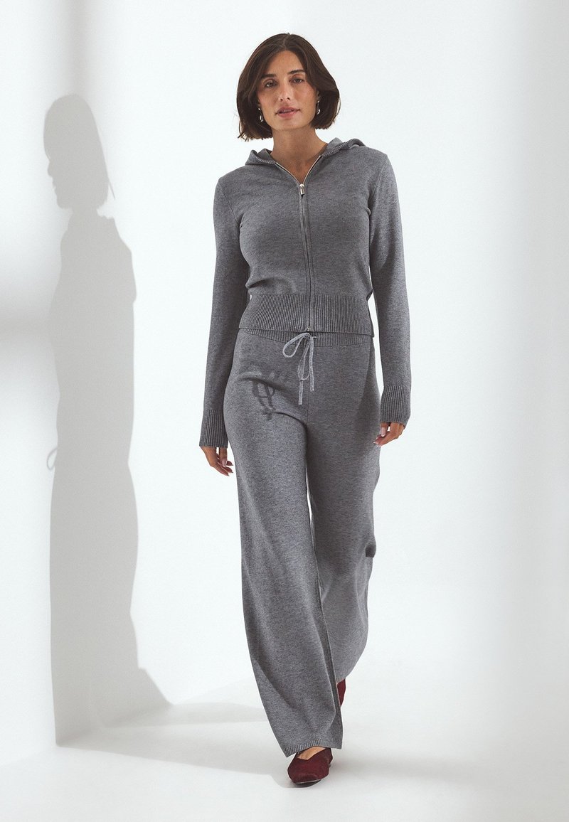 Donna che cammina indossando una felpa grigia con zip e pantaloni a gamba larga abbinati, completati da scarpe rosso scuro, su sfondo bianco uniforme.