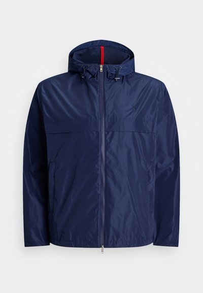 Marinblå lätt jacka med huva, dragkedja fram, två sidofickor, snörningsdetaljer och kontrasterande röd foder vid halsen.