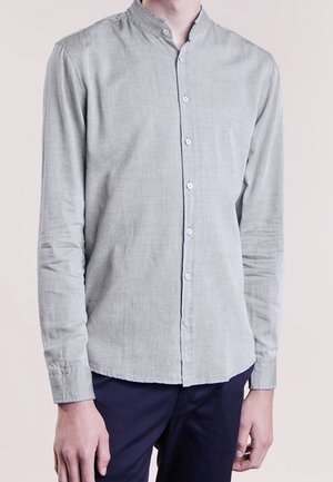 Homme portant une chemise gris clair à manches longues avec un col mandarin et un pantalon bleu marine foncé, debout devant un fond uni.