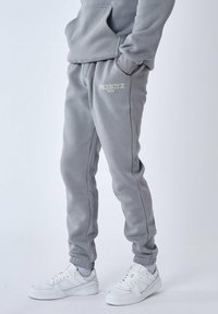 Sudadera de algodón gris con bolsillo frontal y cordón, combinada con pantalones de chándal a juego que tienen rayas laterales y un logo bordado. Zapatillas blancas.