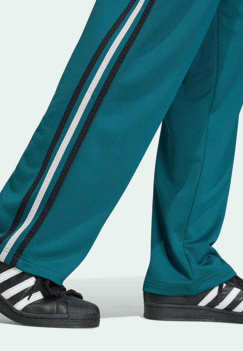 Türkisfarbene Sporthose mit schwarzen und weißen Seitenstreifen, getragen mit schwarzen Sneakern mit weißen Streifen, gezeigt vom Unterschenkel bis zum Fuß.