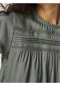 Blouse grise en tissu lisse, avec des détails en dentelle, des plis sur la poitrine et un col rond orné de pompons.