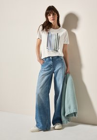 T-shirt gris clair avec un design de texte coloré, associé à un jean bleu délavé à jambes larges. Tenant un pull texturé en vert menthe. Baskets blanches.