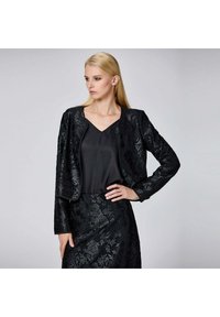 Femme aux cheveux blonds portant une veste texturée noire à motifs floraux et une jupe assortie, associée à un haut noir uni à décolleté en V, posant avec une main sur la hanche.