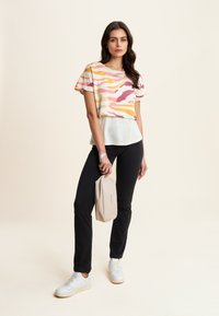 T-shirt multicolore a maniche corte con un motivo a onde, indossato sopra un fondo bianco, abbinato a pantaloni neri e sneakers bianche.