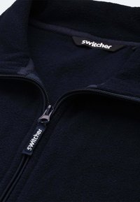 Marineblauer Fleece-Zip-Jacke mit hohem Kragen, ausgestattet mit einem schwarzen Reißverschluss und einem Etikett mit dem "Switcher"-Logo im inneren Kragen. Weiche Textur.