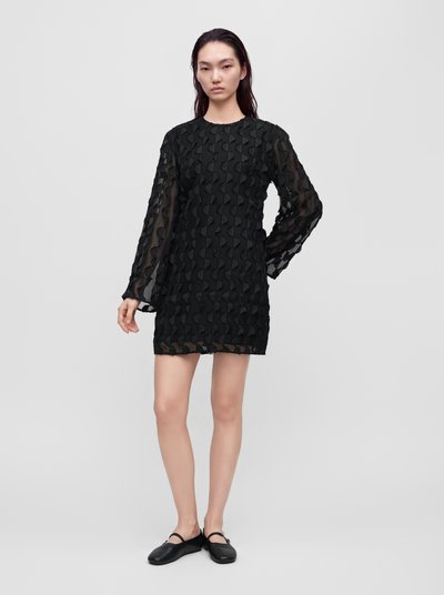 Le mannequin se tient devant un fond de studio gris clair, vêtu d’une mini-robe noire texturée à manches longues et de chaussures plates noires.