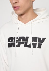 Hoodie aus weißem Stoff mit einem großen schwarzen "REPLAY"-Logo über der Brust. Verstellbare Kordeln und eine weiche Textur.