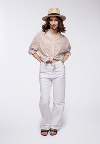 Camicia a righe beige e bianca annodata in vita, abbinata a pantaloni bianchi a gamba larga e sandali neri. Ambientato su uno sfondo chiaro.