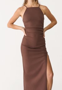 Brun midi-kjole med en tætsiddende silhuet, tynde stropper, slidser i siden og rynkede detaljer langs taljen, lavet af et glat, strækbart stof.