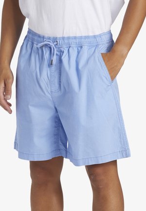 Lyseblå shorts med snøre, båret med en hvid top, den ene hånd i lommen, vist fra midt på låret til taljen på en person med solbrun hud.