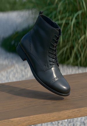 Botín de cuero negro hasta el tobillo con punta redondeada, intrincados detalles de brogue y diseño de cordones. Acabado brillante con un tacón apilado.
