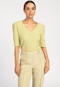 Lichtgele, gestructureerde top met een V-hals en puffed mouwen, gecombineerd met beige hoge taille broeken, weergegeven tegen een effen achtergrond.