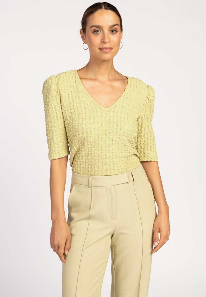 Lichtgele, gestructureerde top met een V-hals en puffed mouwen, gecombineerd met beige hoge taille broeken, weergegeven tegen een effen achtergrond.