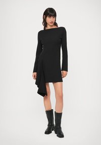 Vestido preto de manga comprida com bainha assimétrica, confeccionado em tecido liso, apresentando decote canoa e um elegante drapeado. São usados botins pretos até ao tornozelo.