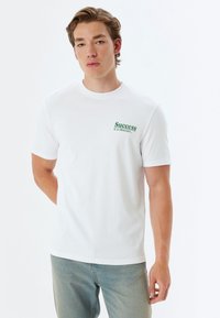 Hvid bomulds T-shirt med rund halsudskæring og korte ærmer, med grøn tekst der står "Succes er en beslutning" på venstre side.