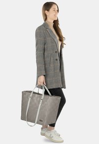 Graue gewebte Tragetasche mit hellgrauen Griffen, getragen von einer Person in einem karierten Blazer und schwarzer Hose, vor einem schlichten weißen Hintergrund.