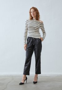 Stripet off-shoulder topp i krem og svart, paret med svarte fuskeskinnbukser og svarte høyhælte sko, stående mot en ensfarget bakgrunn.