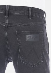 Černé džíny z denimu s hladkou texturou, na zadní kapse obsahující obdélníkovou koženou nášivku s nápisem "Wrangler" a kontrastním prošíváním.