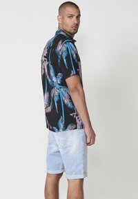 Camisa negra de manga corta con estampado de hojas de palma en rosa y azul, combinada con pantalones cortos de mezclilla claros. La camisa tiene un cuello abotonado y un corte relajado.