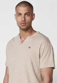 Beige tričká s krátkym rukávom a výstrihom do V vyrobené z jemného materiálu; má malú námornícku logu na ľavej hrudi a voľný strih.