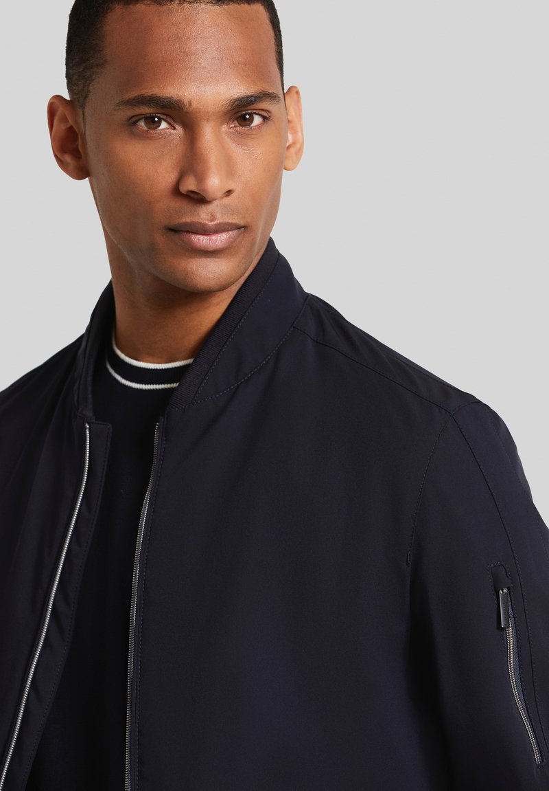 Veste de bombardier navy en tissu lisse, dotée d'une fermeture éclair à l'avant, d'un col côtelé et d'une poche zippée sur la manche.