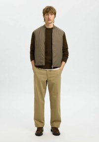 Hellbraune gepolsterte Weste über einem dunkelbraunen Pullover, beigen Hosen und dunkelbraunen Schuhen; stehend auf einem schlichten Hintergrund.