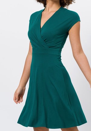 Mujer con un vestido cruzado de manga corta en un tono teal profundo, con cintura ajustada y falda acampanada, vista desde el cuello hasta la rodilla sobre un fondo liso.