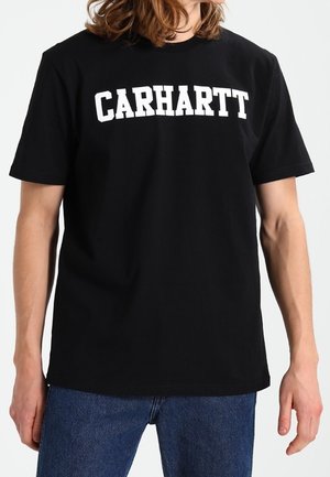 Osoba nosząca czarną koszulkę Carhartt z białym logo i niebieskie dżinsy, stojąca na białym tle.
